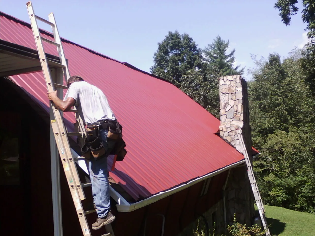 Roofer on a ladder beside a red metal roof and stone chimney — Soffit & Fascia Repair by Roofing Brothers in Center City